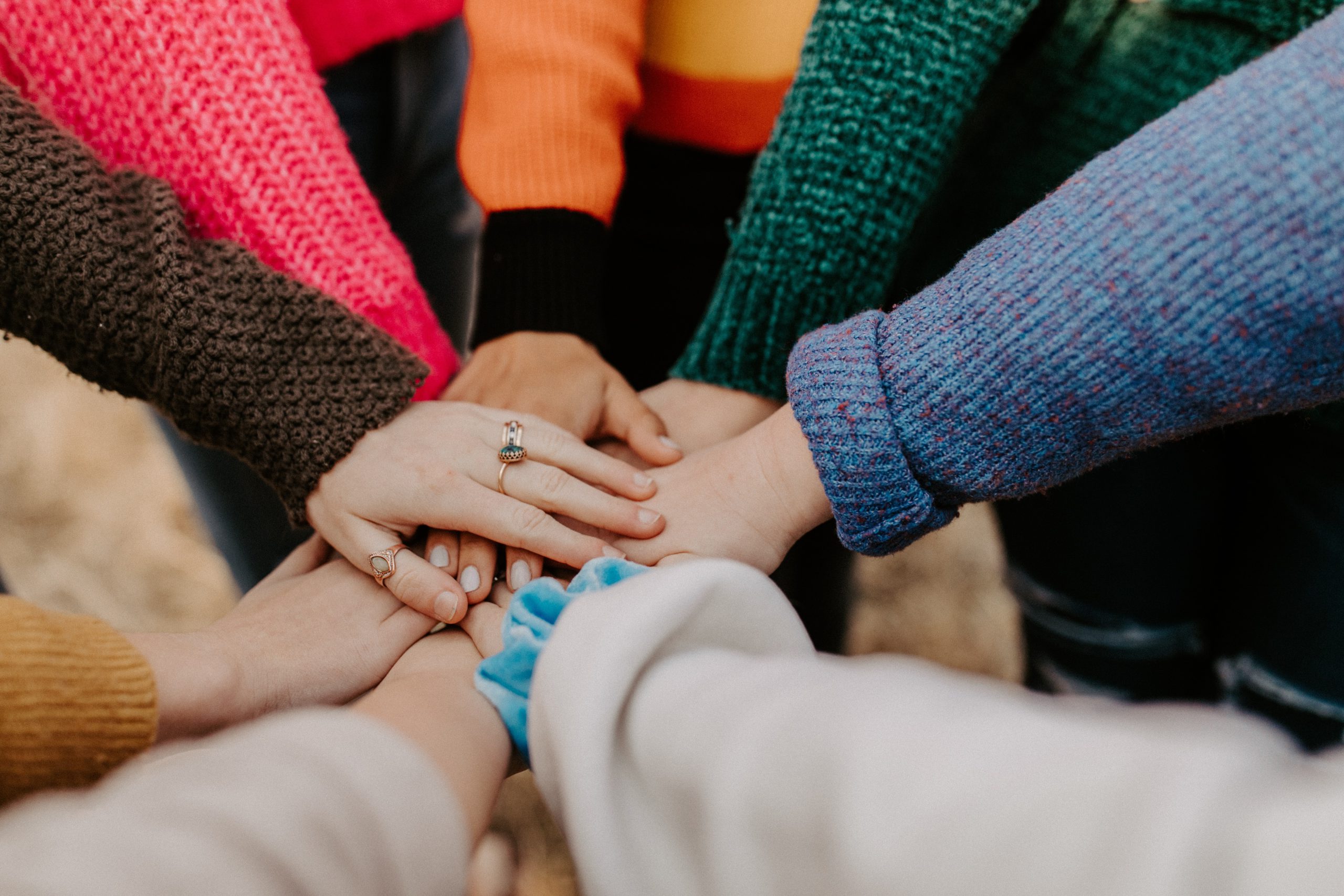 People wearing bright colored long sleeve sweaters are putting their hands together in a circle