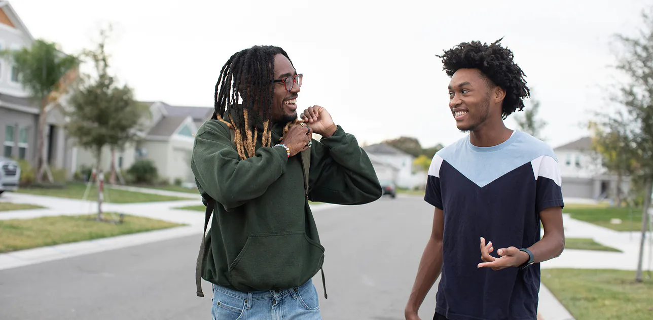 Two people are walking and chatting on a suburban street. They appear relaxed and cheerful, wearing casual clothes. Trees and houses line the street.