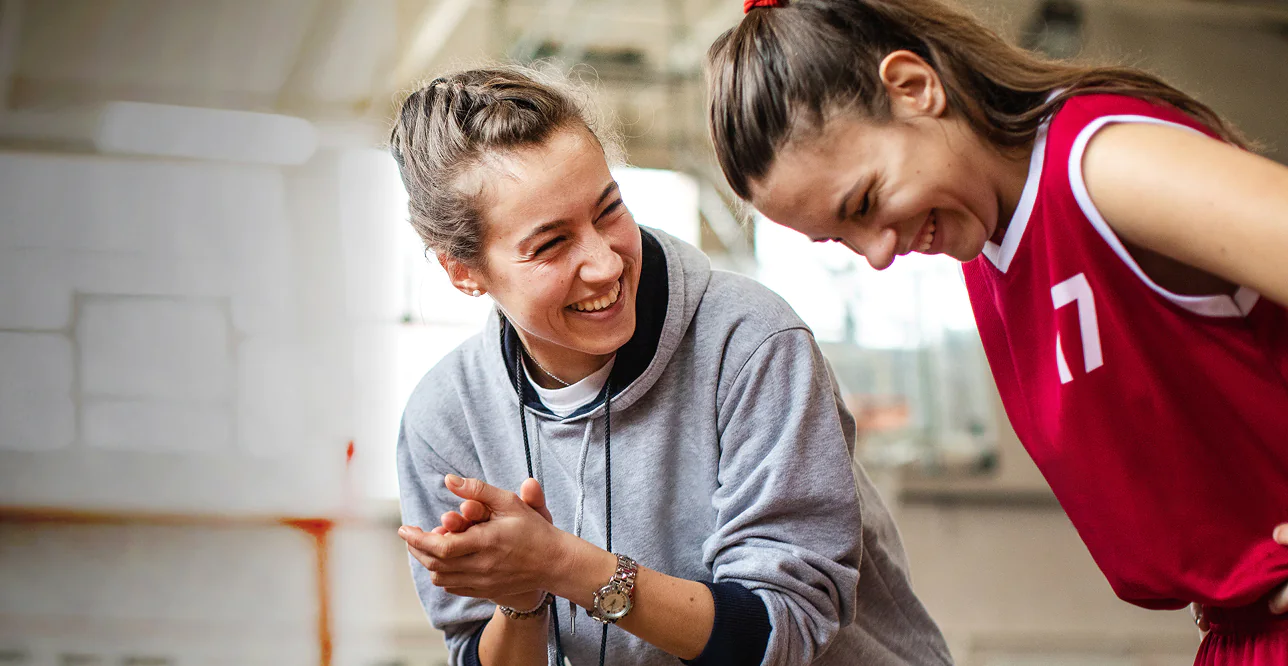 Two women share a joyful moment in a sports setting. One wears a gray hoodie, clapping, while the other in a red jersey leans in, both smiling brightly.